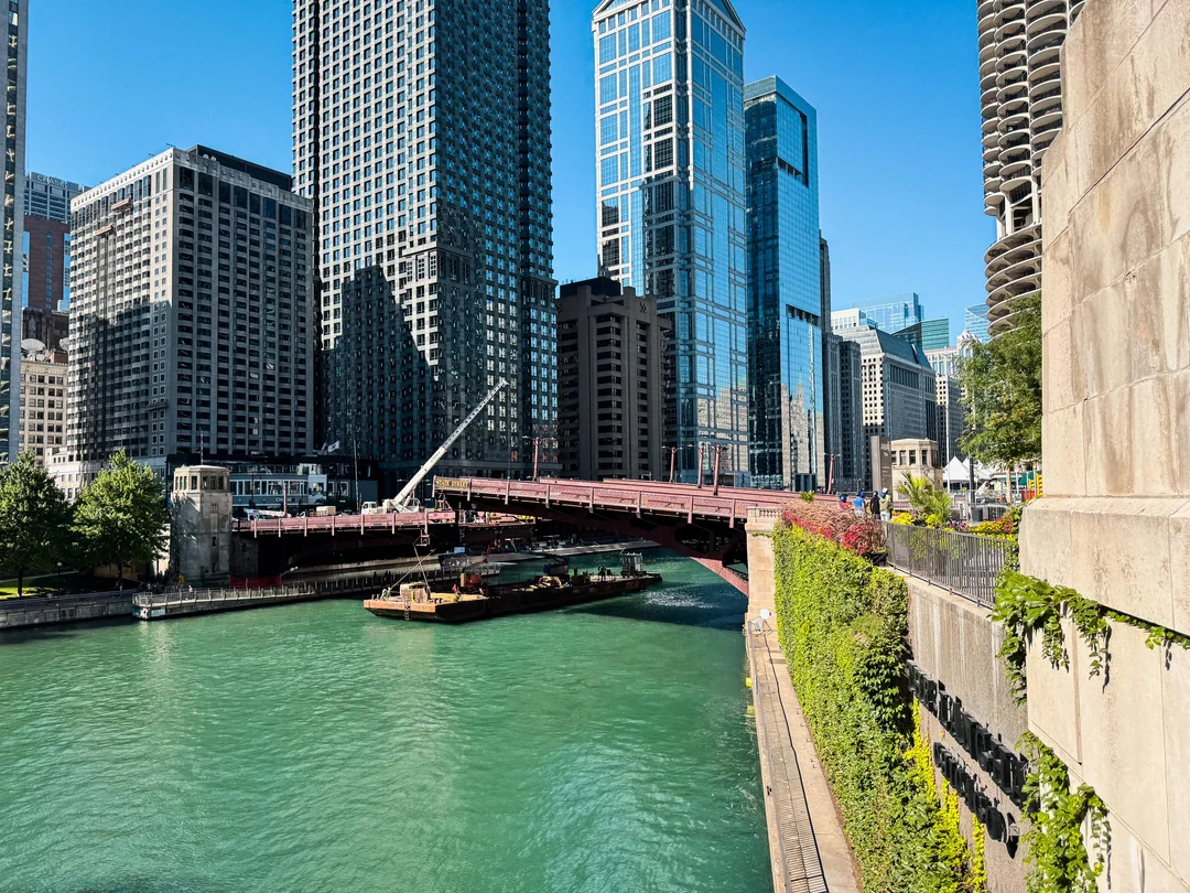 A river with turquoise water runs under a red bridge in a cityscape of tall, modern glass buildings. A barge and crane are on the water, with greenery along the riverbank and bright sunlight illuminating the scene.
