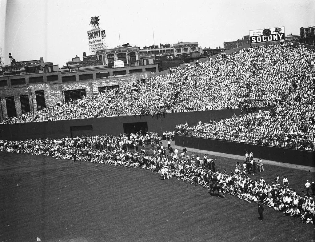 Black-and-white photo of a crowded baseball stadium with fans seated in the stands and on the field. Large SOCONY ads are visible on the outfield wall and rooftop in the background.