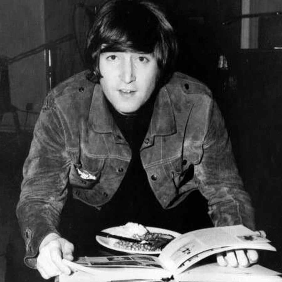 A person with medium-length dark hair wearing a jacket looks up while holding a book open, with a plate of food on the table in front of them, in a black and white photo.