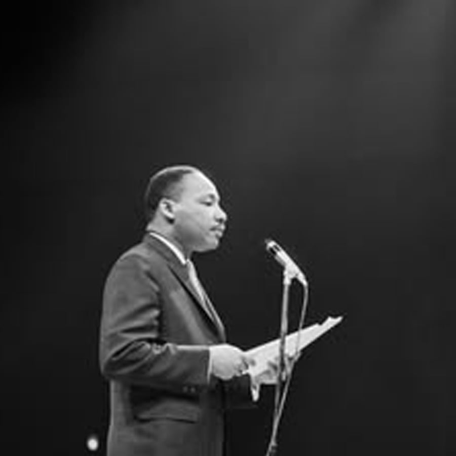 A man in a suit stands at a microphone, holding papers and speaking under a spotlight, with a dark background behind him.
