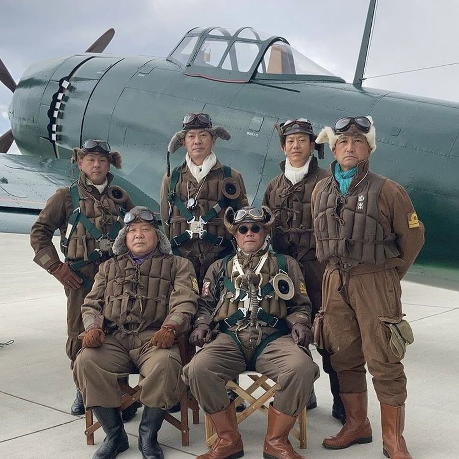 Six people in vintage pilot uniforms pose in front of a green World War II-era fighter plane on a runway. Four stand in the back, two sit in front, all wearing goggles and brown flight suits.