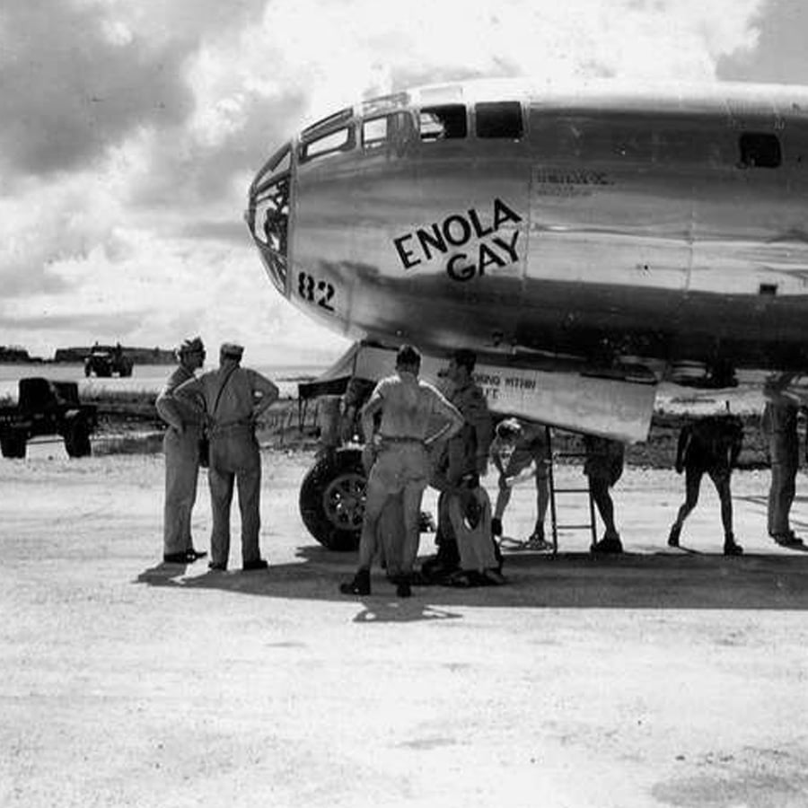 A group of military personnel stand near the nose of the Enola Gay, a large military aircraft, on an airfield under a cloudy sky.