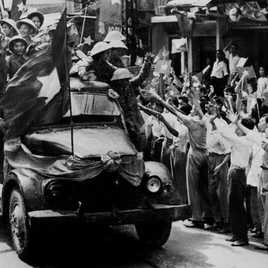 Soldiers on a military vehicle holding flags are greeted by a crowd of cheering people on a busy street, some reaching out their hands. The scene appears to be a historical celebration or parade.