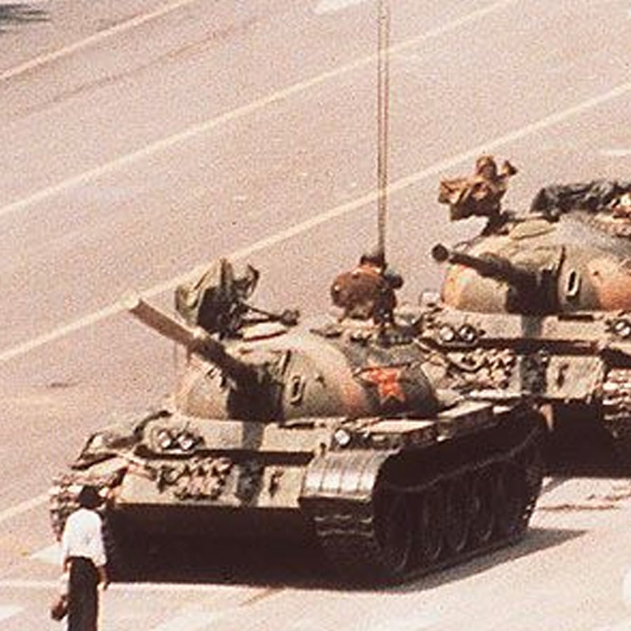 A person stands in front of a line of military tanks on a wide city street, blocking their path. The scene is in daylight, and the tanks appear to be stopping for the individual.