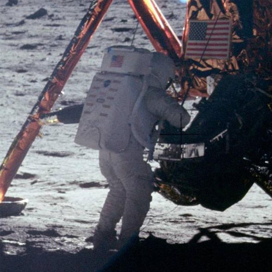 An astronaut in a spacesuit stands next to a lunar module on the Moon’s surface, with the American flag visible on both the module and the suit. The ground appears dusty and rocky under a dark sky.