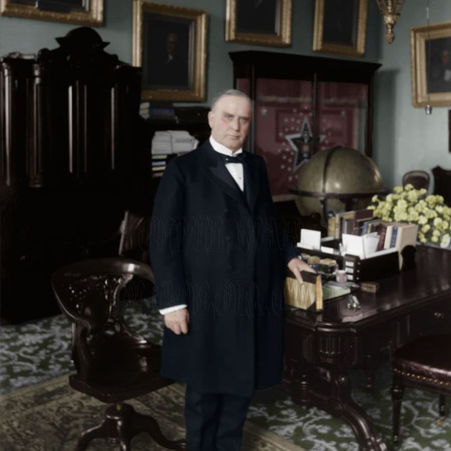 A man in a dark suit stands beside an ornate desk in a formal office with framed portraits, a globe, and a vase of yellow flowers in the background.