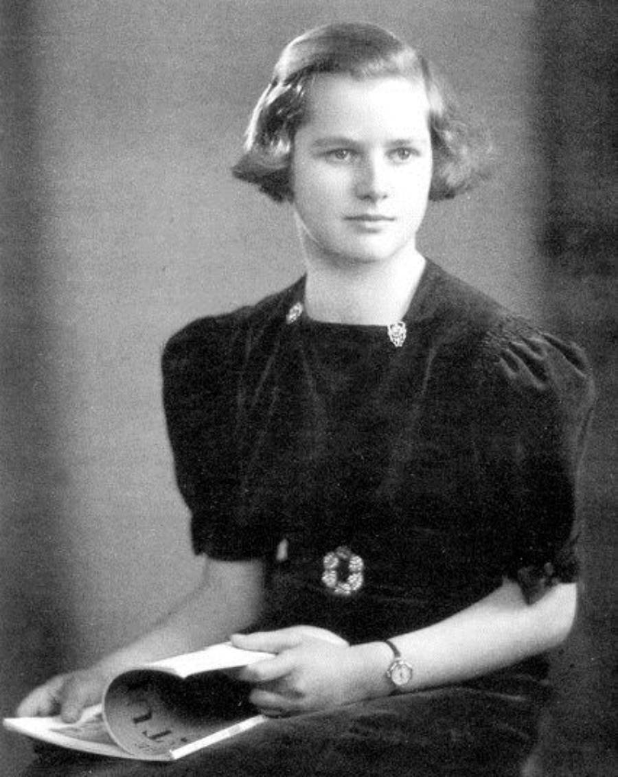 A young woman with short hair, wearing a dark dress, sits holding an open magazine or book. She looks slightly to the side with a calm expression in this black-and-white portrait photograph.