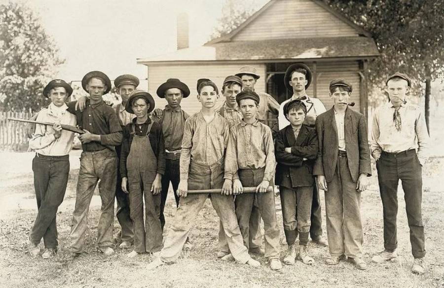 Thirteen boys in old-fashioned clothing pose outdoors in front of a house, some holding sticks or bats, with serious and neutral expressions. Trees and a fence are visible in the background.
