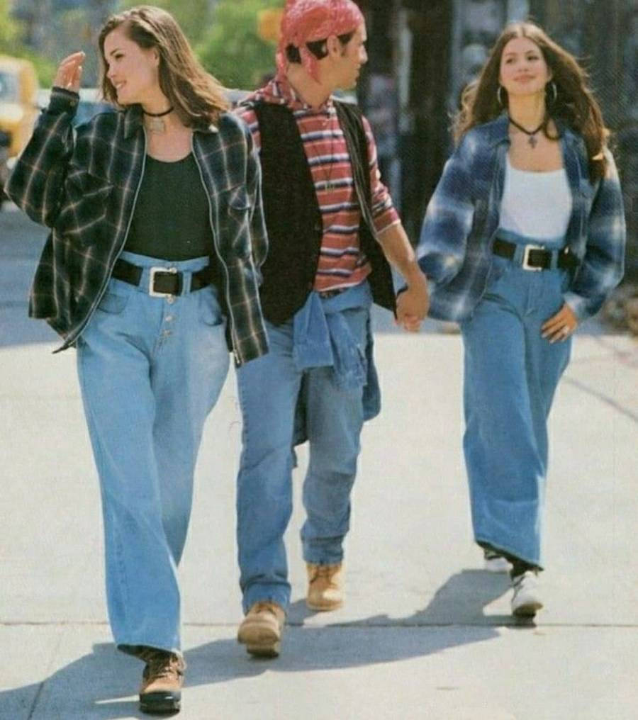 Three young adults walk on a sidewalk. Two women wear loose jeans, black tops, and plaid shirts, while the man between them wears a striped shirt, jeans, a vest, and a red bandana. They appear to be enjoying a casual day out.