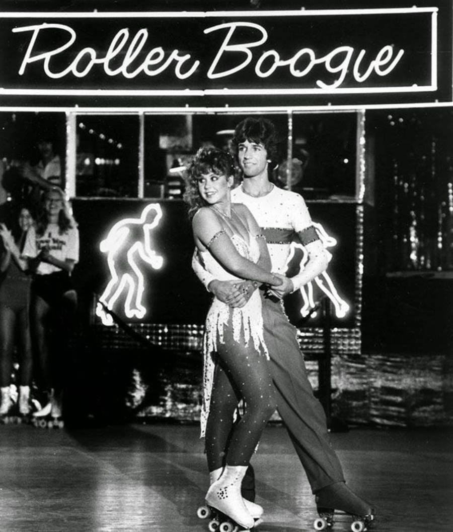 A couple in sparkly outfits roller skate together in front of a neon “Roller Boogie” sign, with silhouettes of skaters and people watching in the background.
