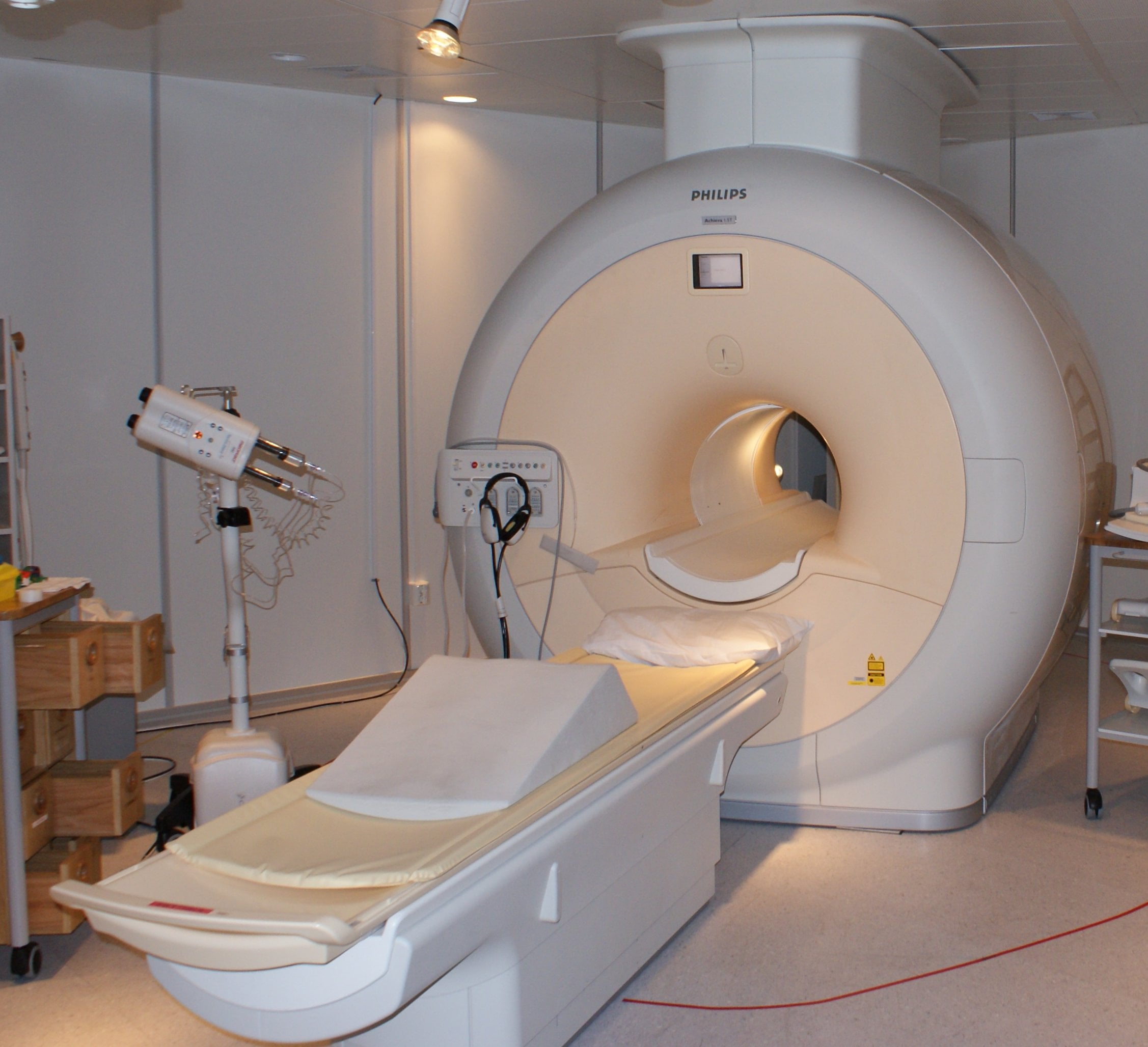 An MRI machine in a hospital room with a padded table for patients to lie on and medical equipment nearby. The scanner has a large circular opening and is labeled Philips.