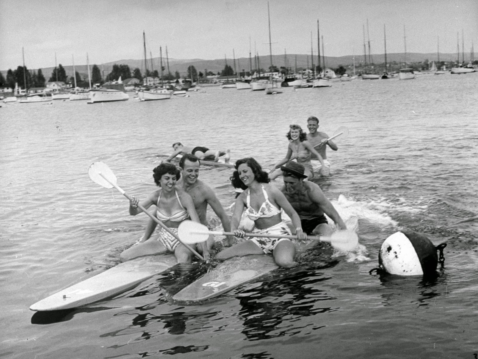 Four women and two men in swimsuits playfully paddleboard and splash in a harbor with boats and yachts in the background. A buoy floats nearby; everyone looks happy and energetic, enjoying a sunny day on the water.