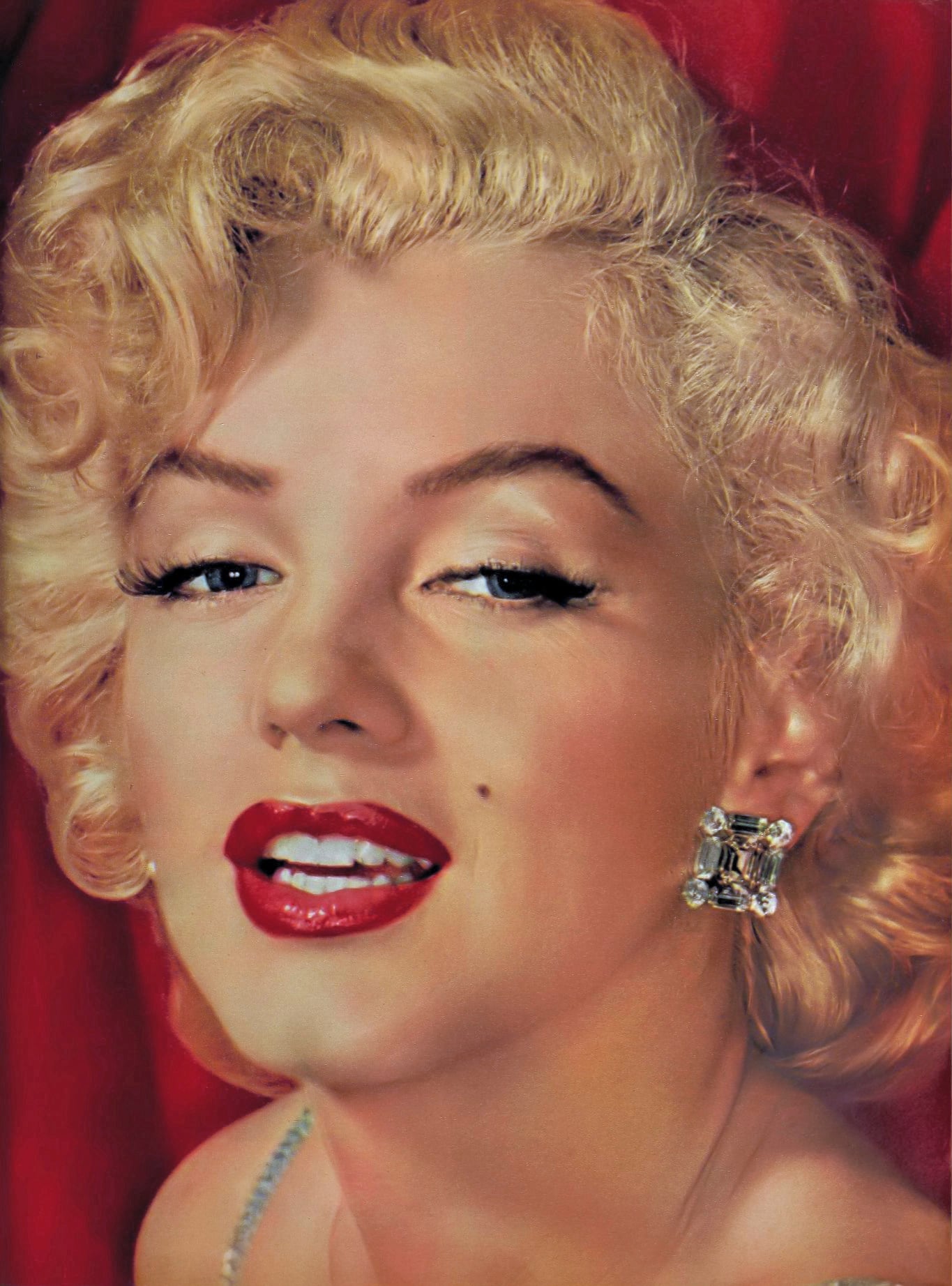 A woman with short, curly blonde hair, wearing bold red lipstick, dark eyeliner, and a large square earring, smiles slightly in front of a red background.