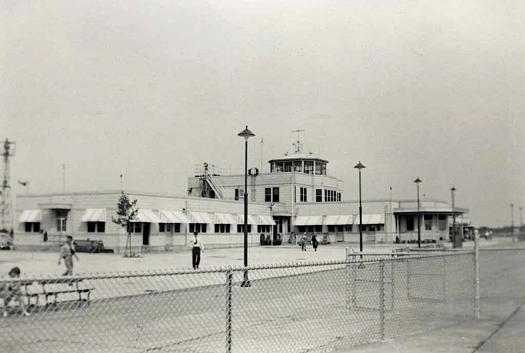 Black and white photo of an airport terminal with a control tower, people walking, benches, lamp posts, and a chain-link fence in the foreground.