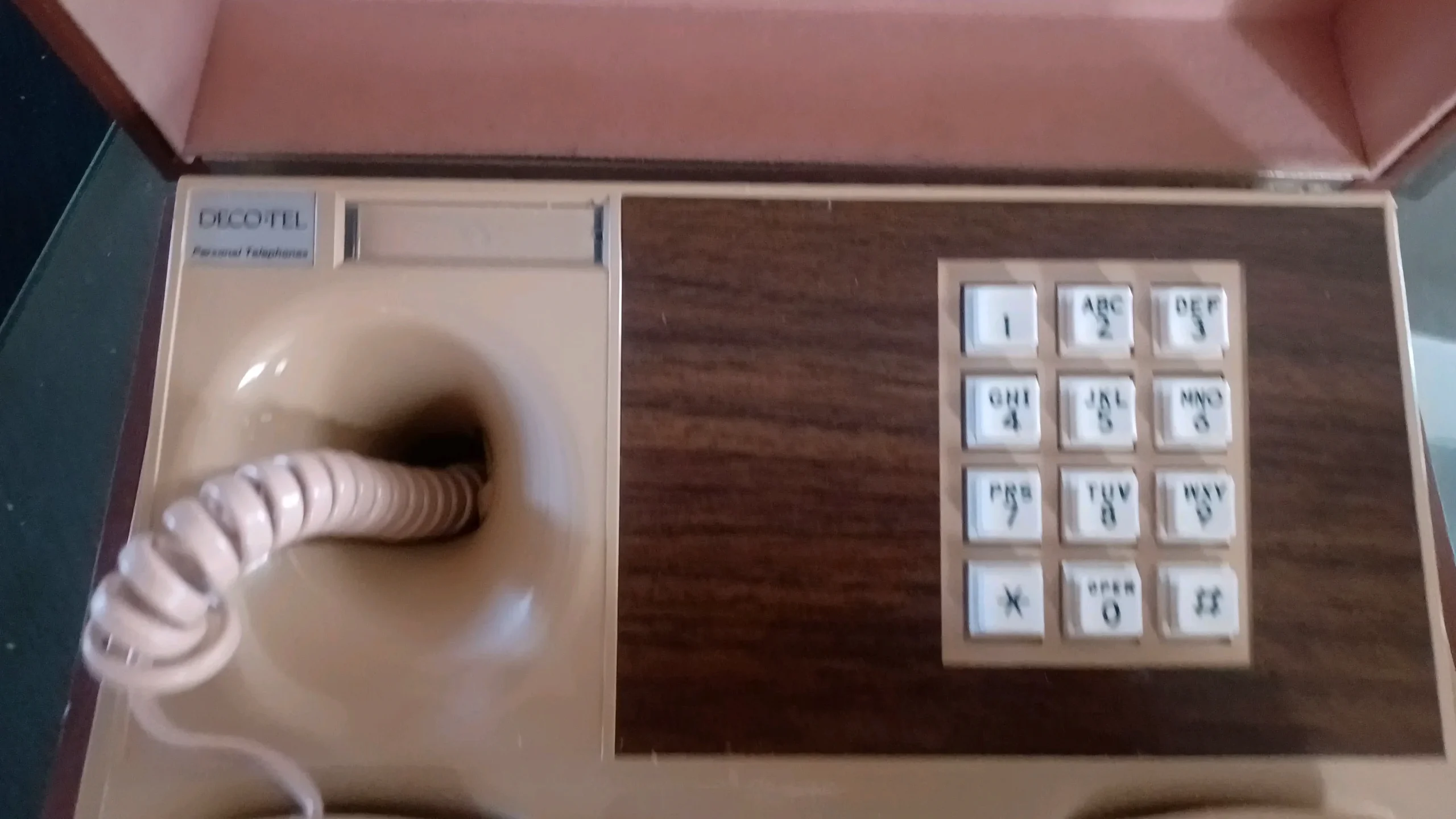 A vintage beige and brown push-button telephone with a coiled cord, woodgrain panel, and labeled keys. The brand “Decotel” is visible in the upper left corner.