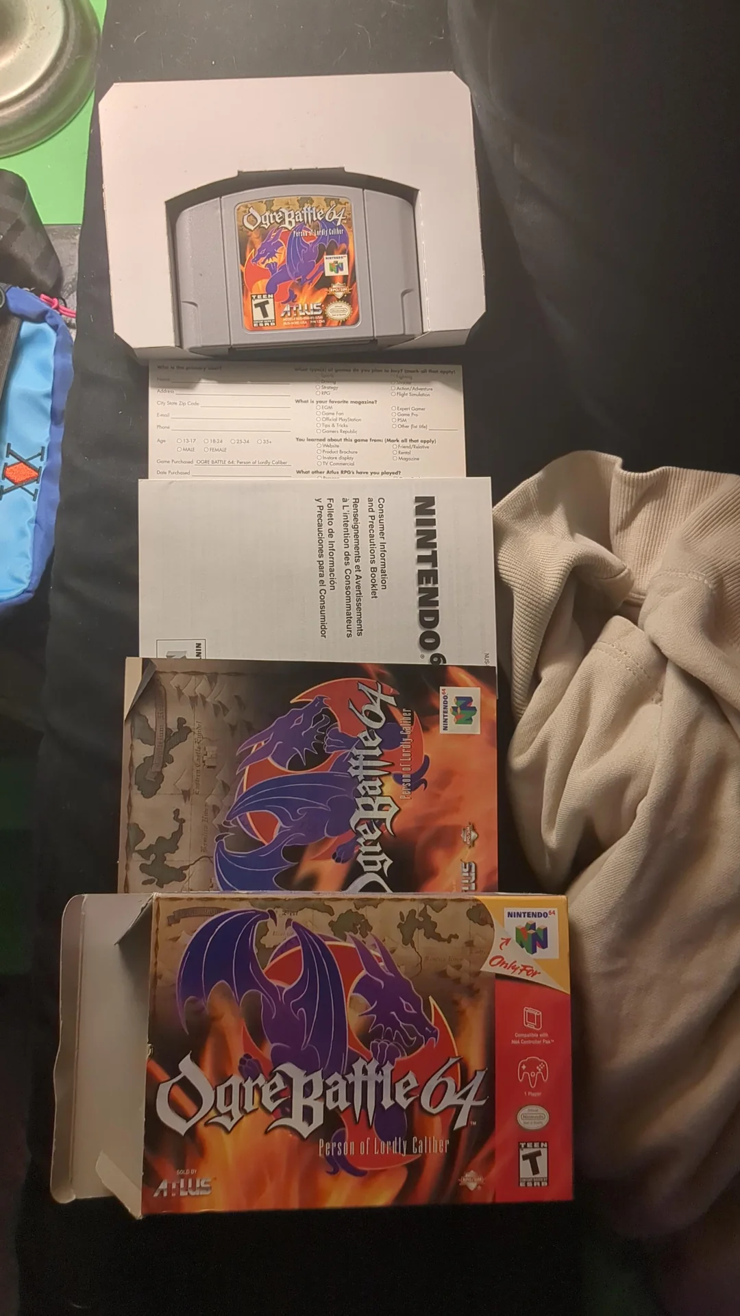 Ogre Battle 64 Nintendo 64 game cartridge, manual, and box lay on a surface with a blanket and blue bag nearby. The box features a dragon and flames design.