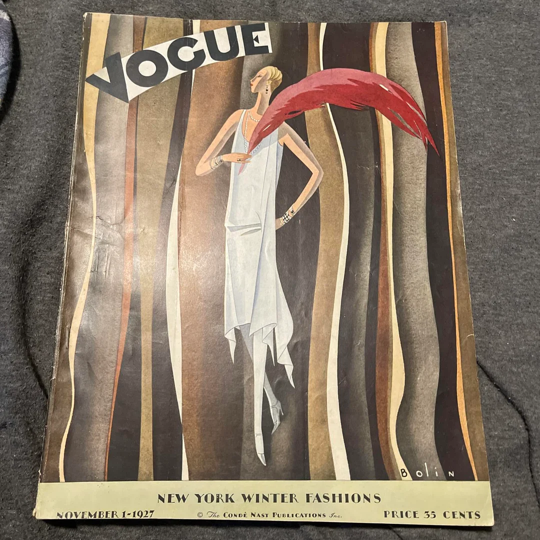 Vintage Vogue magazine cover from November 1, 1927. An illustrated woman in a white dress with a red feather stands against an abstract striped background. Text below reads: “New York Winter Fashions.”