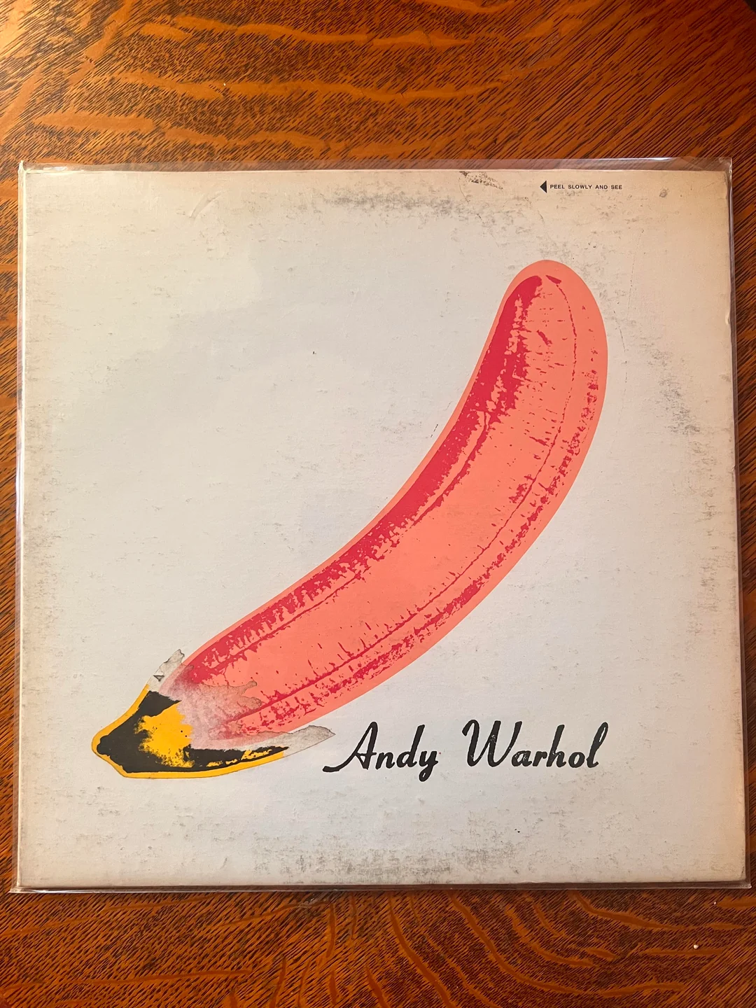 A vinyl album cover featuring a large pink banana illustration on a white background, with “Andy Warhol” written in black cursive at the bottom right. The cover sits on a wood surface.