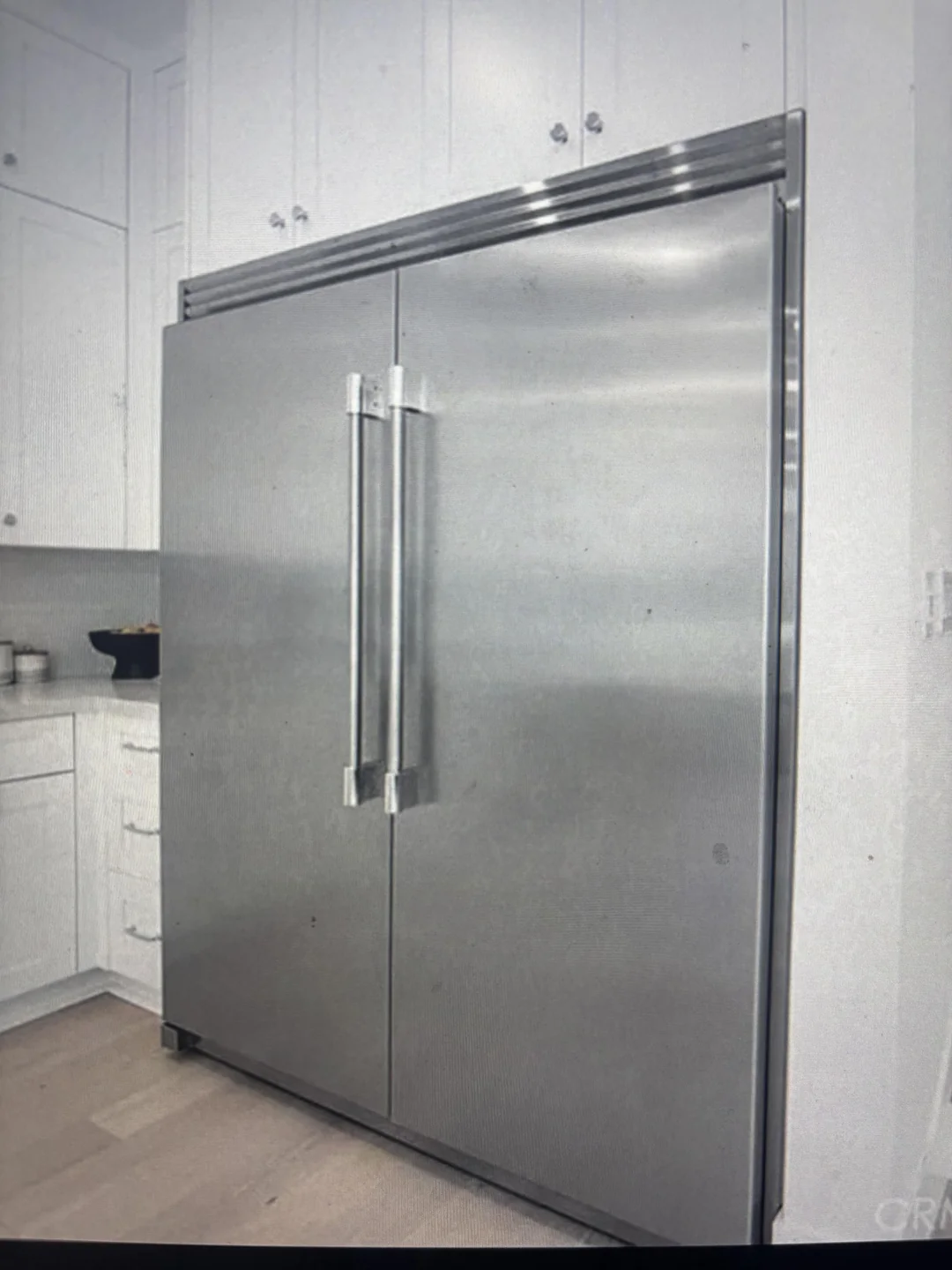 A large stainless steel double-door refrigerator is built into a white kitchen with cabinets and drawers around it. The floor is light wood.