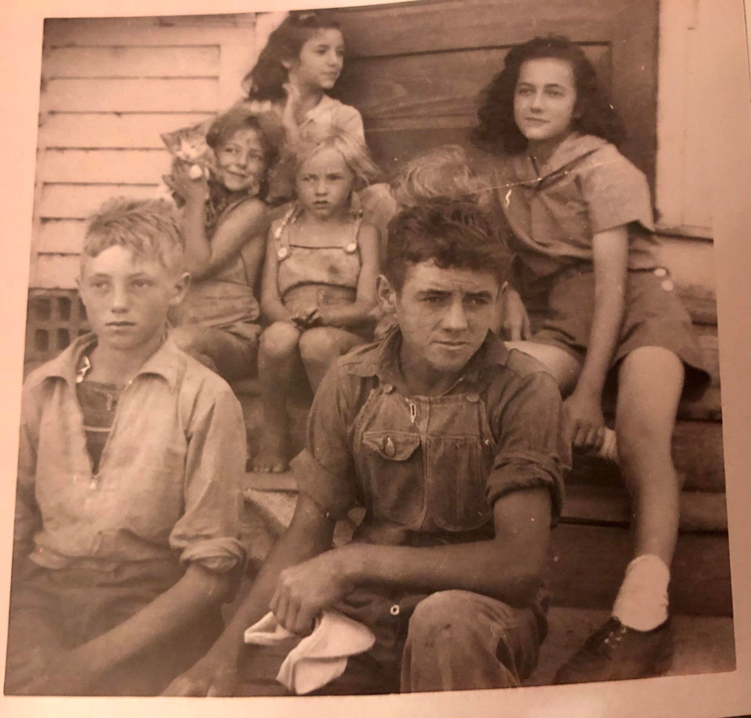 Six children sit and pose on outdoor wooden steps. Three girls are on the top step, two boys and one girl sit in front. The children wear casual clothing and look at the camera. The photo is in black and white.
