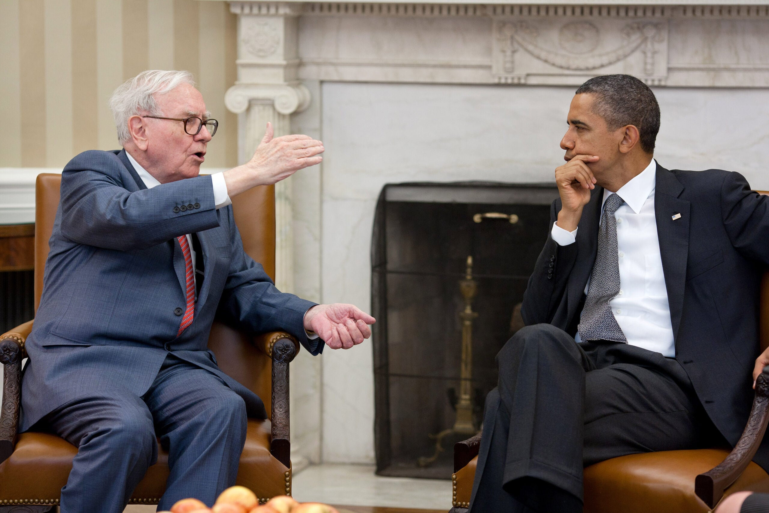 Two men in suits sit in armchairs, engaged in conversation in a formal office with a fireplace in the background. One gestures while speaking; the other listens thoughtfully. A bowl of apples sits on the table in front.