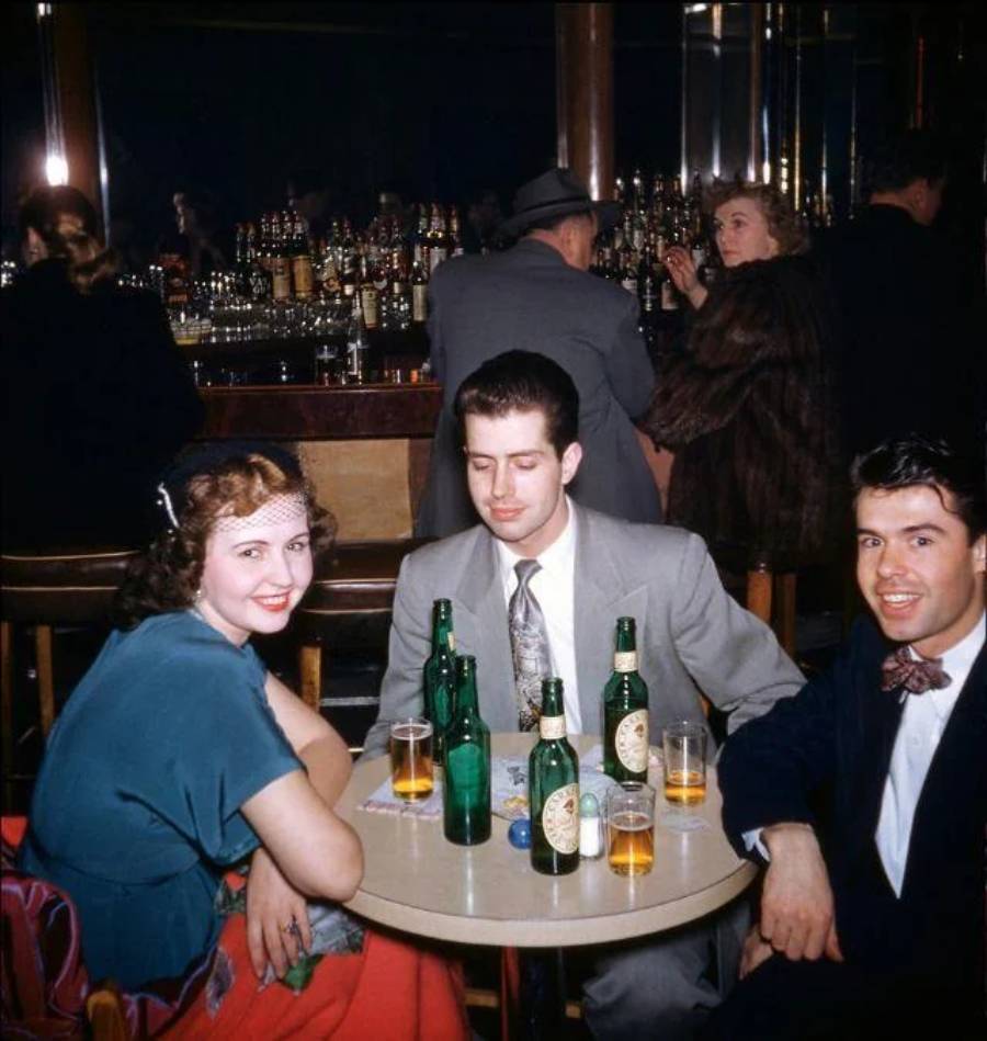Three people sit at a round table with beers and glasses in a lively bar, smiling. The background shows a mirrored wall, bar counter, bottles, and other patrons.