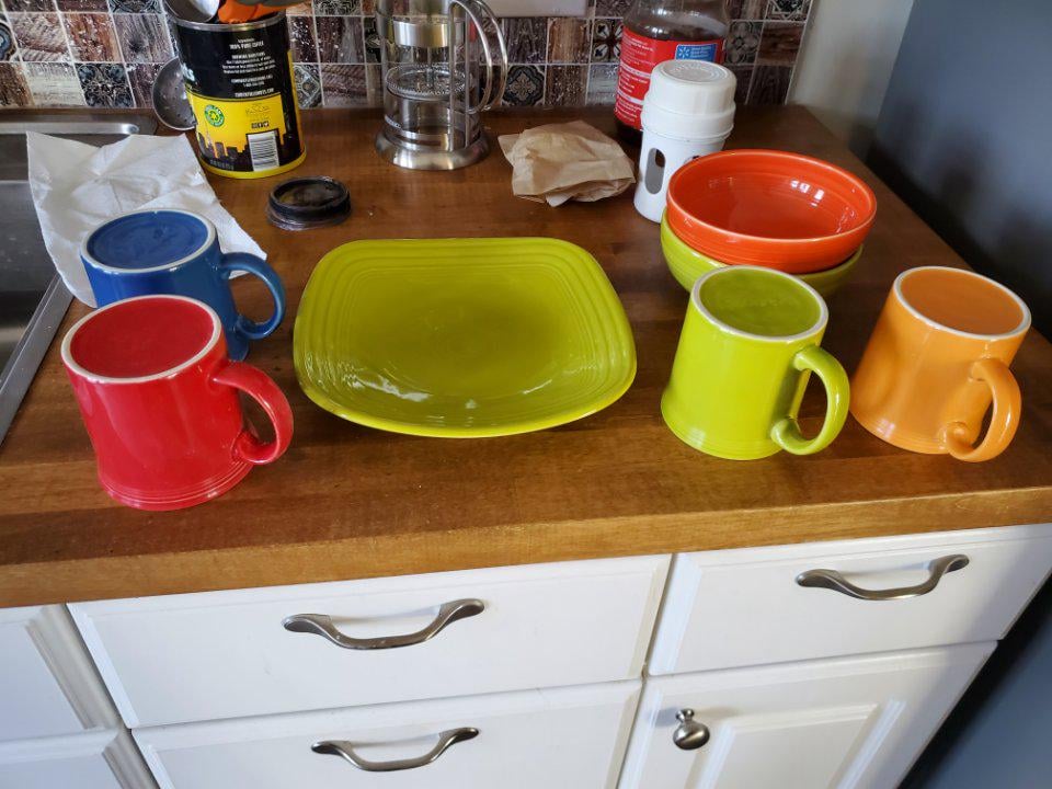 Four colorful mugs, an empty green plate, and three orange bowls are arranged on a wooden kitchen countertop. Various kitchen items and a napkin are also visible in the background.