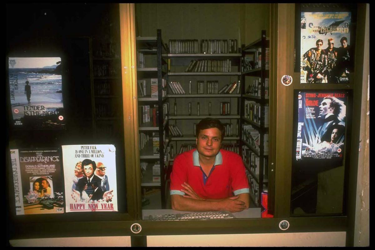 A man in a red shirt sits behind a glass counter in a video rental store, surrounded by movie posters and shelves filled with VHS tapes.
