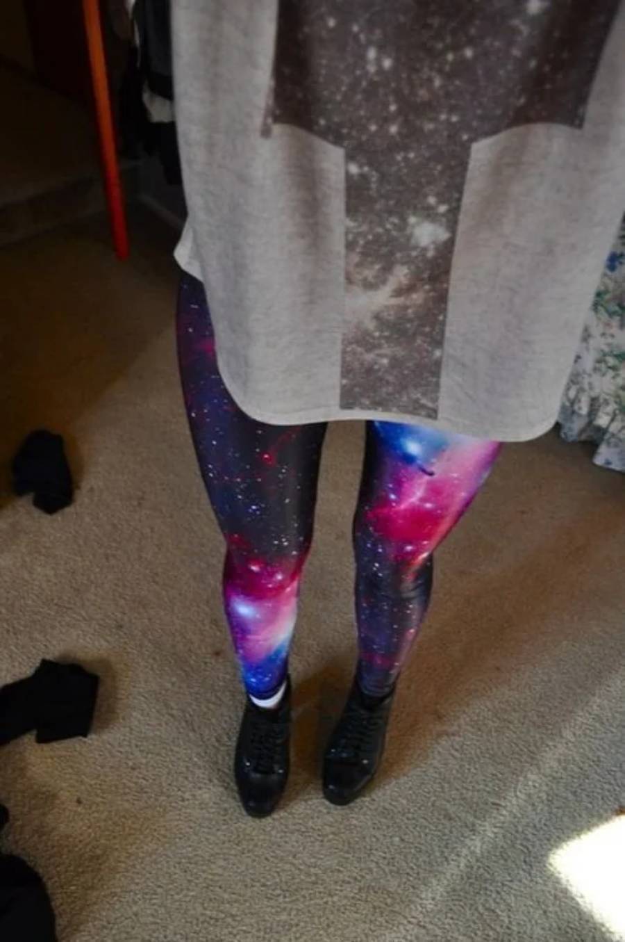 Person wearing galaxy-patterned leggings and black shoes, standing on a beige carpet. Their shirt is partially visible, and clothing items are on the floor nearby.