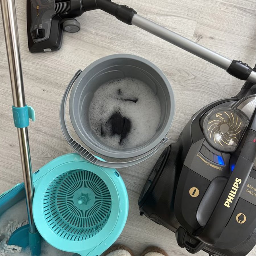 A mop, bucket with soapy water, and a black Philips vacuum cleaner on a light wooden floor, seen from above. Part of a turquoise spin mop set and a pair of white shoes are also visible.