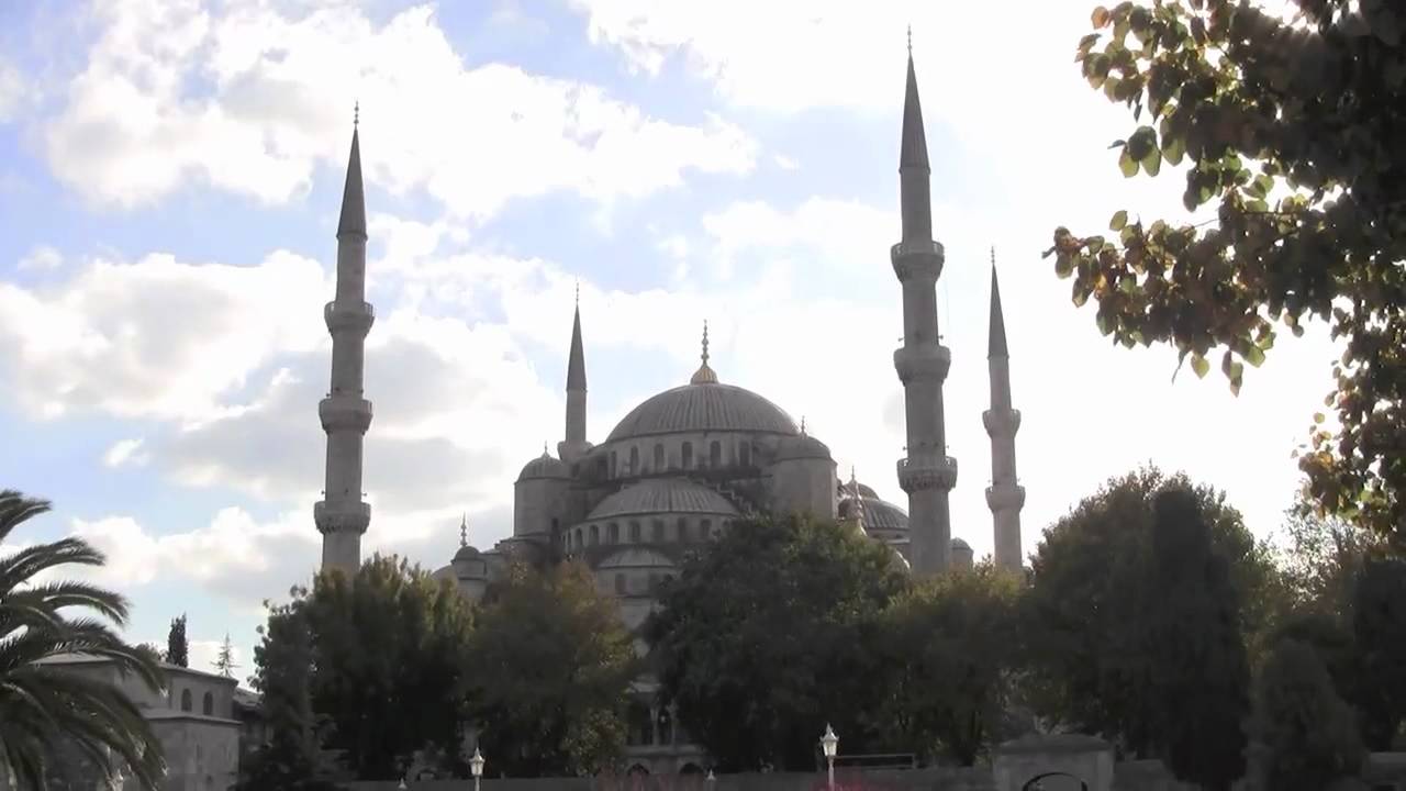 The Blue Mosque in Istanbul, Turkey, with six tall minarets and a large central dome, is silhouetted against a bright sky, surrounded by trees.