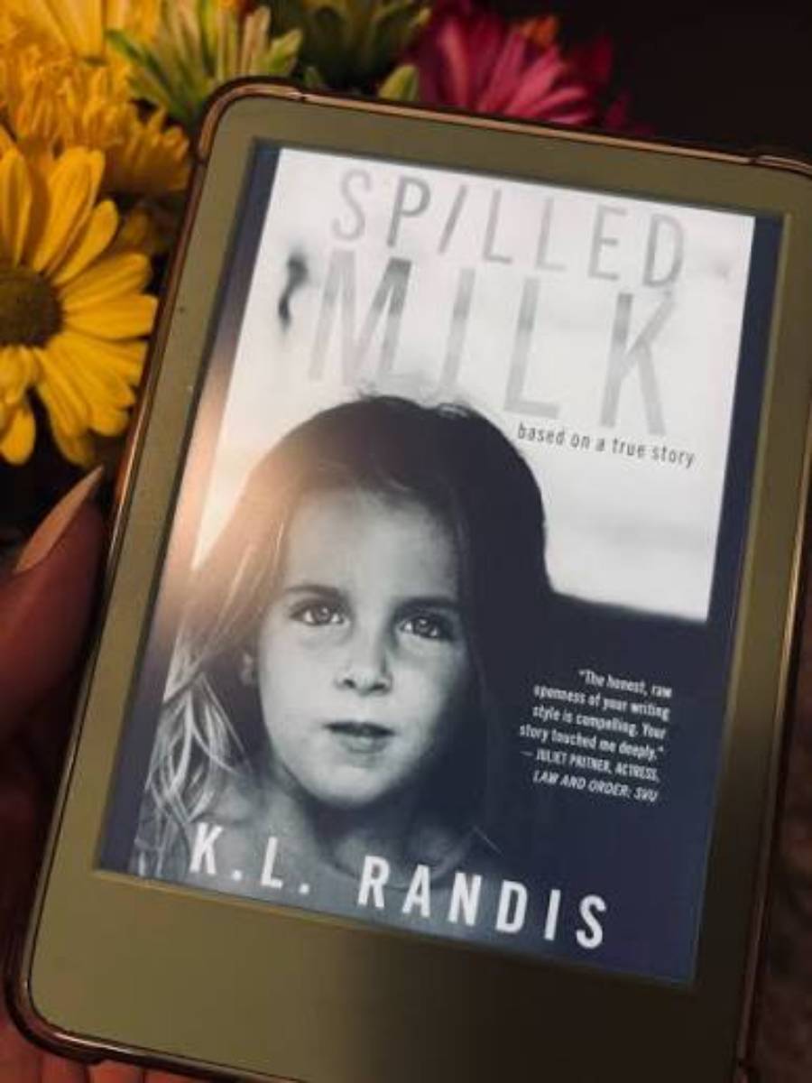 A person holds an e-reader displaying the cover of “Spilled Milk” by K.L. Randis. The cover features a black-and-white photo of a young girl, flowers are visible in the background.