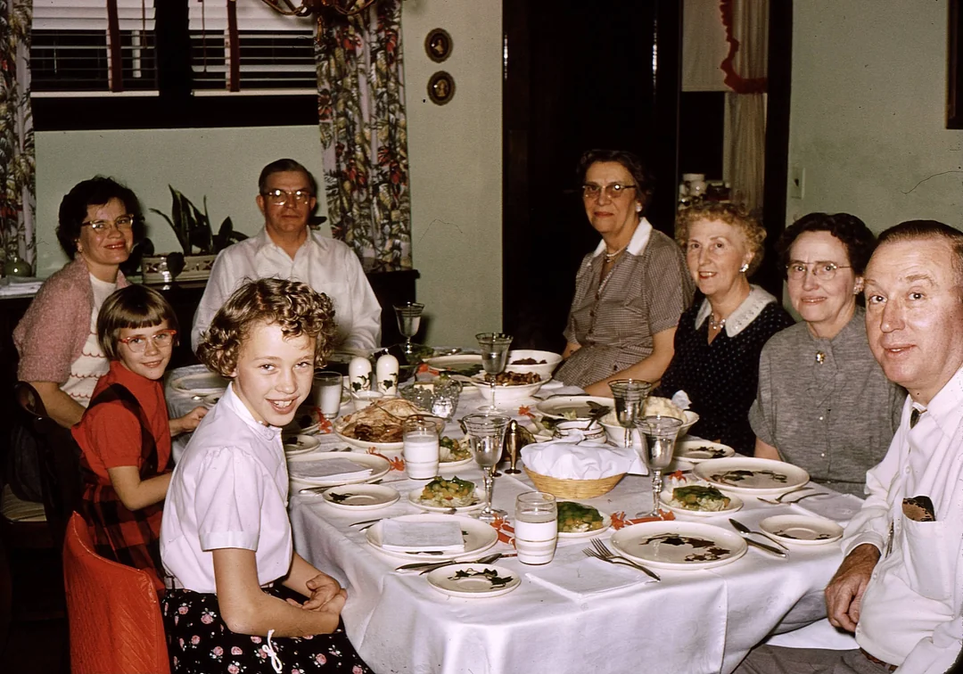 Seven people, including two children, sit around a dining table set for a meal. The table has plates, glasses, and food. The group smiles, appearing to enjoy a family gathering in a warmly decorated room.