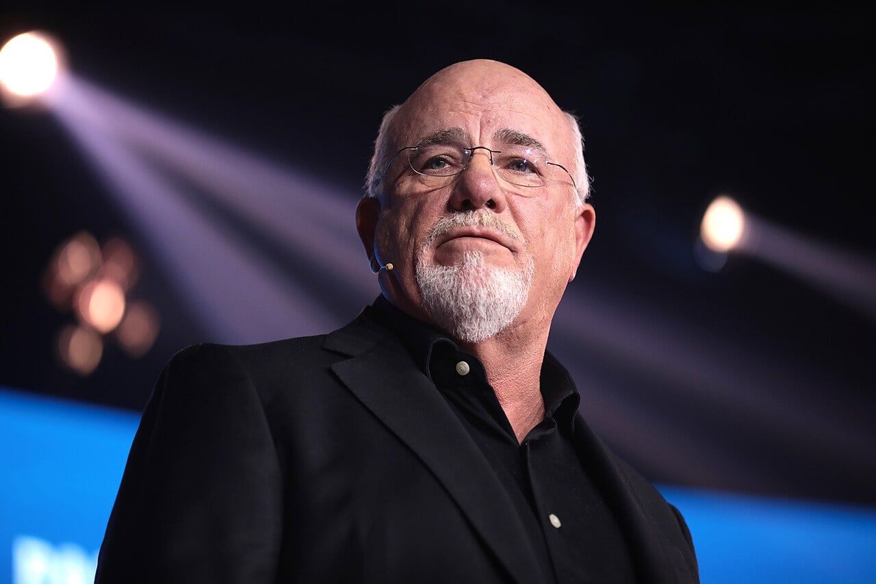A bald man with glasses, a white goatee, and a black suit jacket stands on stage under dramatic lighting, looking thoughtfully into the distance.