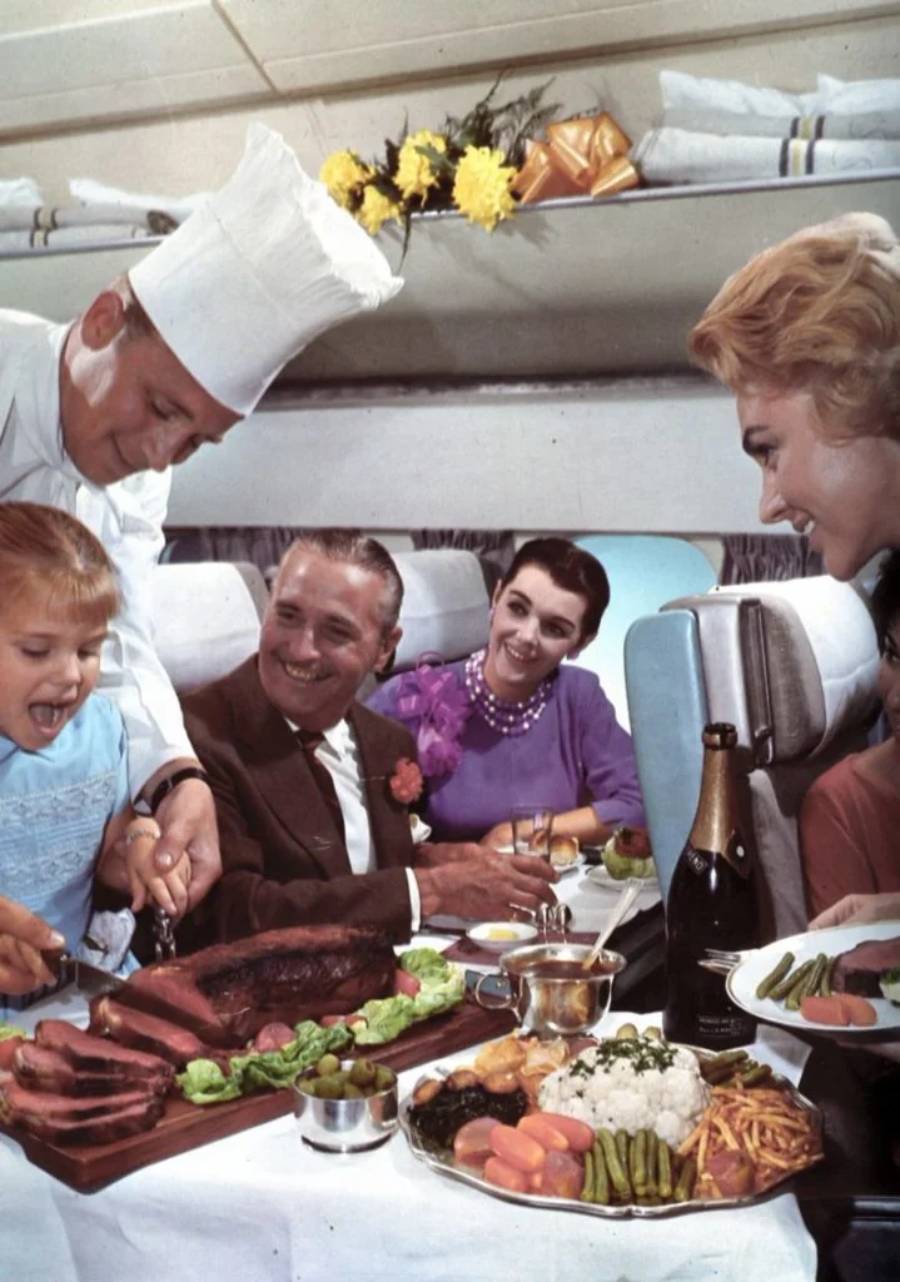 A chef serves steak to a smiling family on an airplane. The table is filled with food, vegetables, and champagne. The atmosphere is elegant and joyful, with everyone dressed formally.