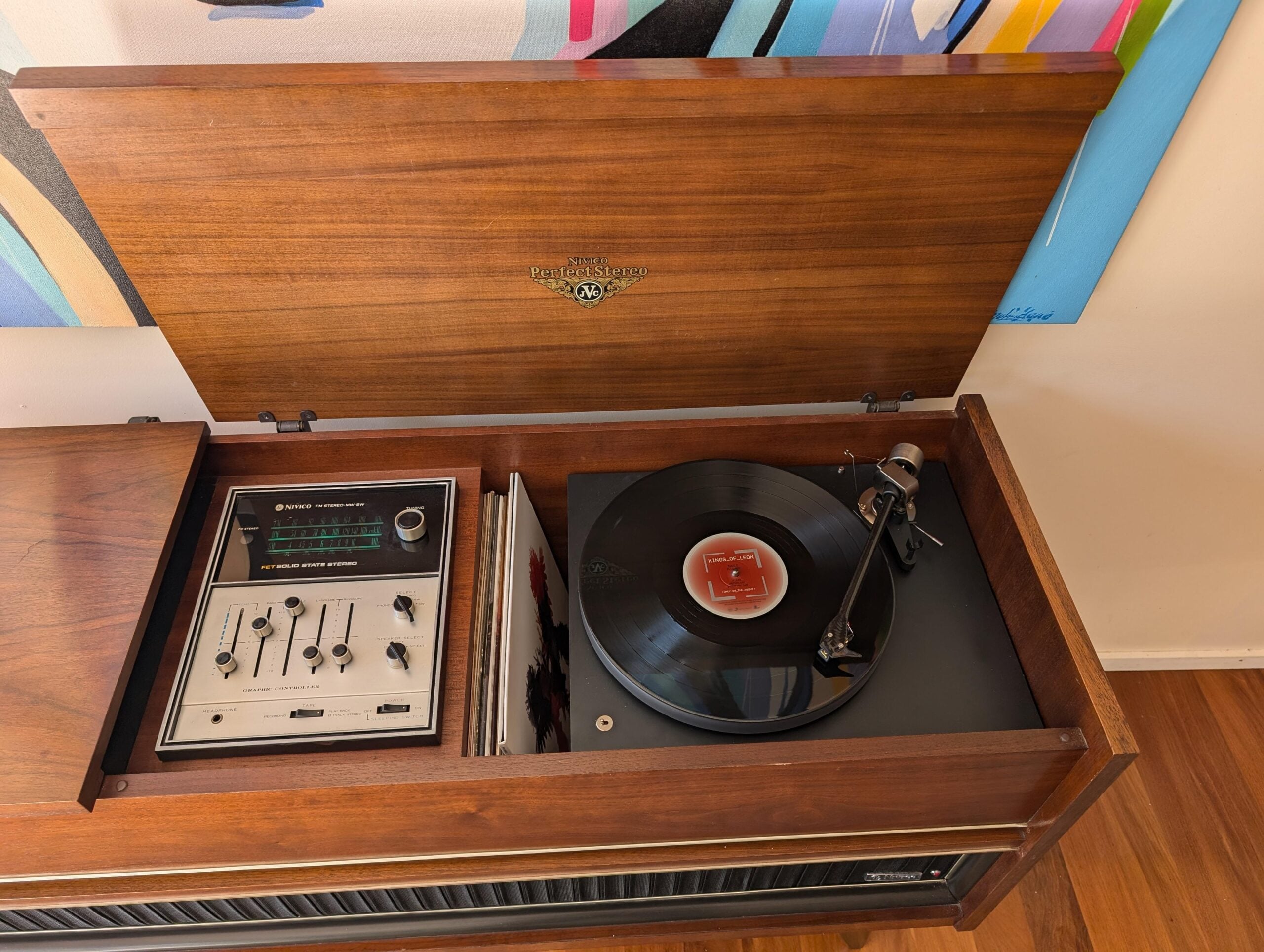 A vintage wooden record player console with a black vinyl record spinning, open lid, control panel with knobs and switches, and colorful artwork partially visible in the background.