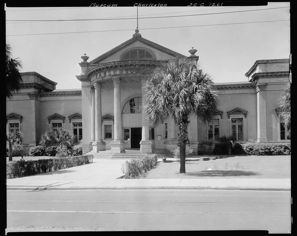 Historic museum building with large columns at the entrance, arched windows, and a triangular pediment. A palm tree and manicured lawn are in front, with a sidewalk and street in the foreground.
