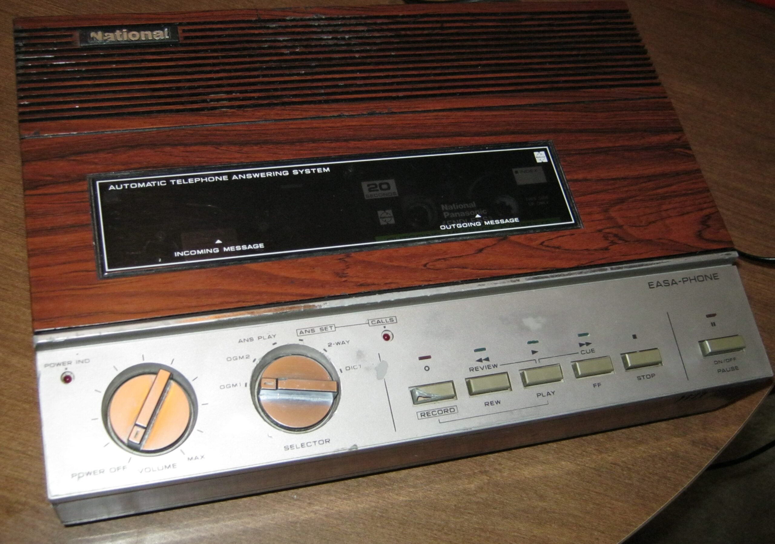 A vintage National Easa-Phone answering machine with a woodgrain finish, buttons, and dials for playback, recording, and message management, sitting on a wooden surface.