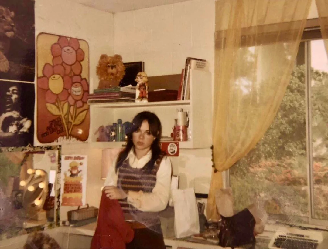 A woman stands in a decorated room near a window with yellow curtains, shelves with toys and books, and colorful posters, including a flower and a musician. She holds a red garment and looks toward the camera.