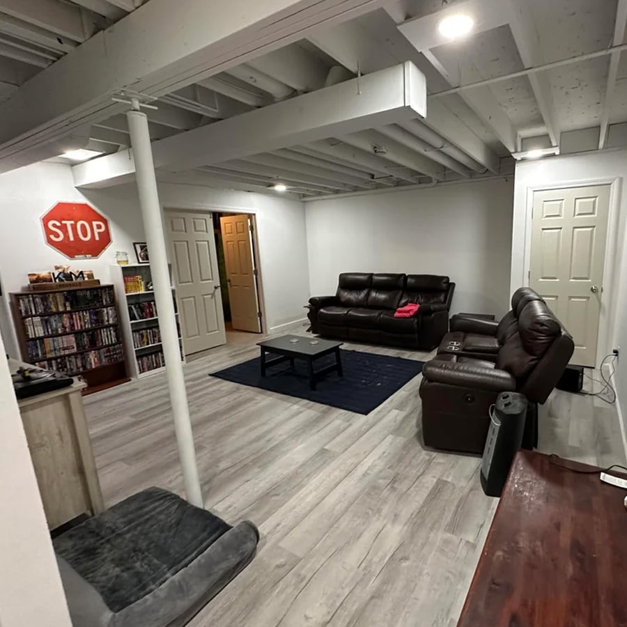 Basement room with exposed ceiling, two brown leather sofas, a small table on a blue rug, shelves of DVDs, a TV stand, and a stop sign on the wall. Multiple doors and wood-style flooring are visible.