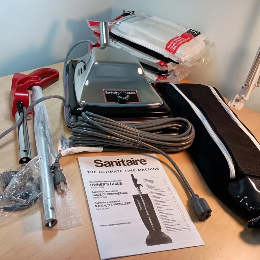 A Sanitaire vacuum cleaner with accessories, hose, owner’s guide, vacuum bags, and black carrying bag arranged on a wooden table.