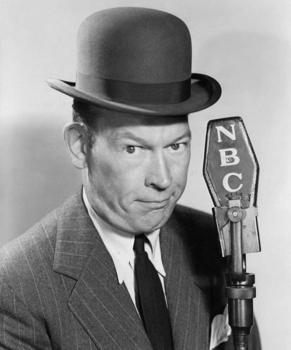 A man in a suit and bowler hat stands next to an old-fashioned NBC microphone, looking slightly amused. The image is in black and white, giving it a vintage feel.