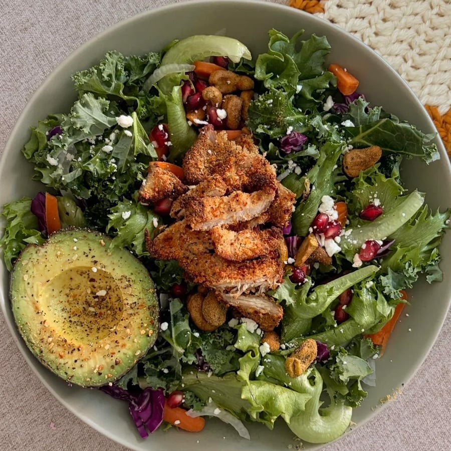 A bowl of mixed greens salad topped with grilled chicken, half an avocado, pomegranate seeds, nuts, feta cheese, and sliced vegetables on a beige tablecloth.
