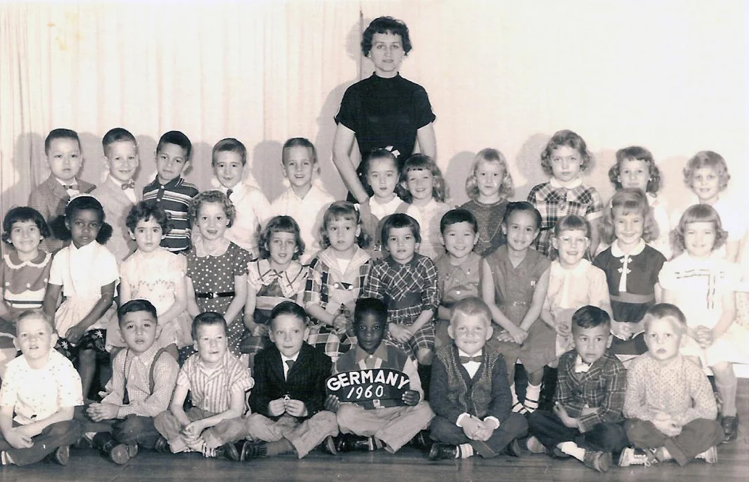 Black and white photo of a teacher standing behind a group of young children posed in three rows; a boy in front holds a sign reading “GERMANY 1960.” The students wear 1960s-style clothing and smile at the camera.