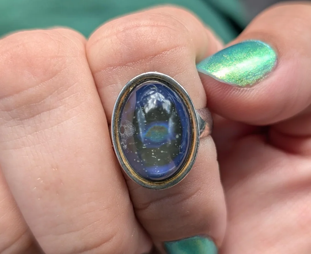 A close-up of a hand with metallic green nails holding a silver ring featuring an oval blue stone, which reflects outdoor scenery and light patterns.