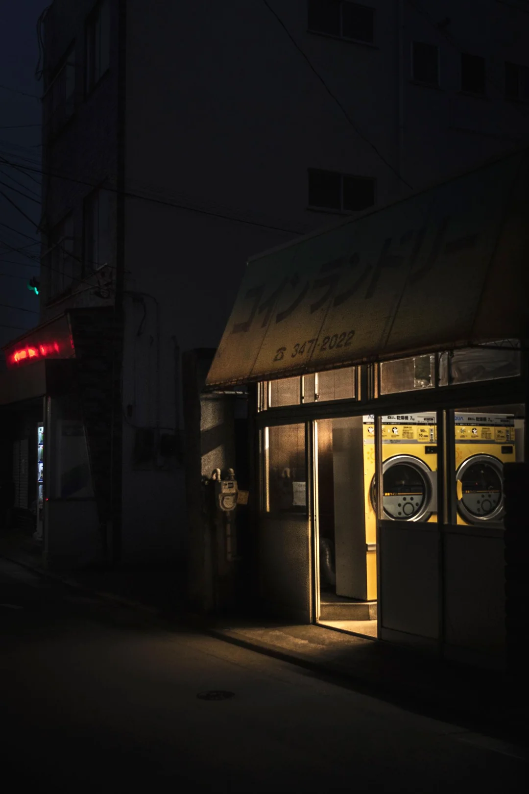 A dimly lit laundromat with yellow washing machines, illuminated at night. The street outside is dark, with only a small neon sign glowing at a nearby building.