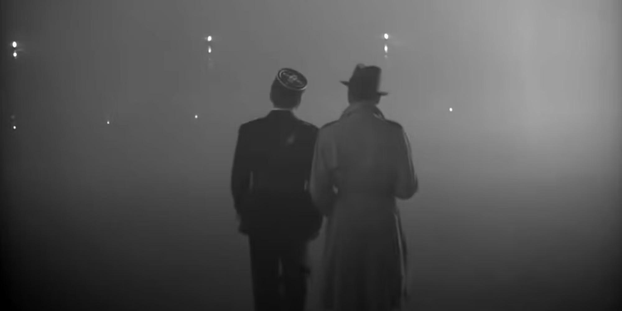 Two people in dark coats and hats walk side by side into heavy fog, with faint lights visible in the distance, in a black-and-white scene.
