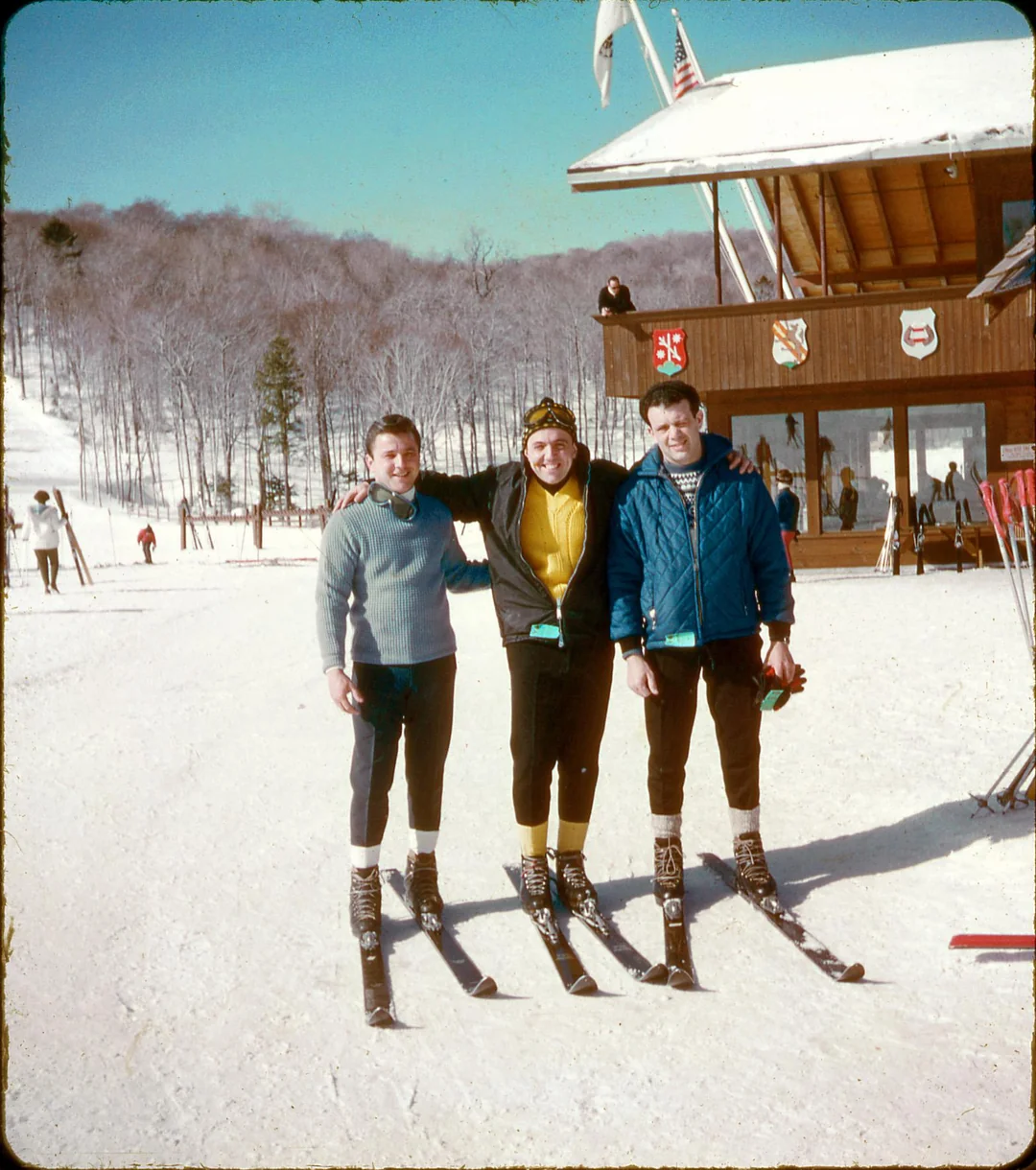 Three people in vintage ski outfits stand smiling on snowy ground near a wooden ski lodge, with leafless trees and bright blue sky in the background. Skis and ski poles are visible in the snow around them.