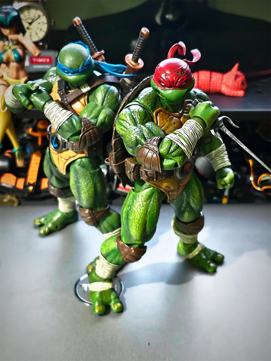 Two Teenage Mutant Ninja Turtles action figures, one with a blue mask and two swords, the other with a red mask and two sais, stand in dynamic poses on a tabletop with blurred objects in the background.