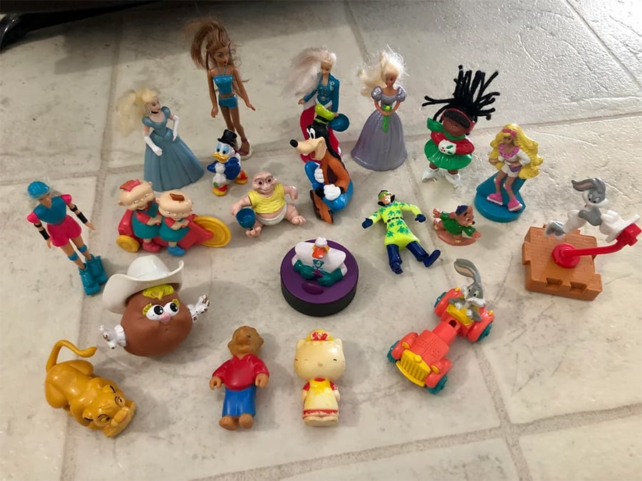A variety of small toys, including action figures, animals, cartoon characters, and vehicles, are scattered on a light-colored tiled floor. The toys are arranged randomly and vary in color, shape, and size.
